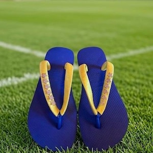 May include: A pair of blue flip-flops with yellow straps, resting on a green grassy field. The straps have a decorative pattern. The flip-flops are a casual footwear choice.