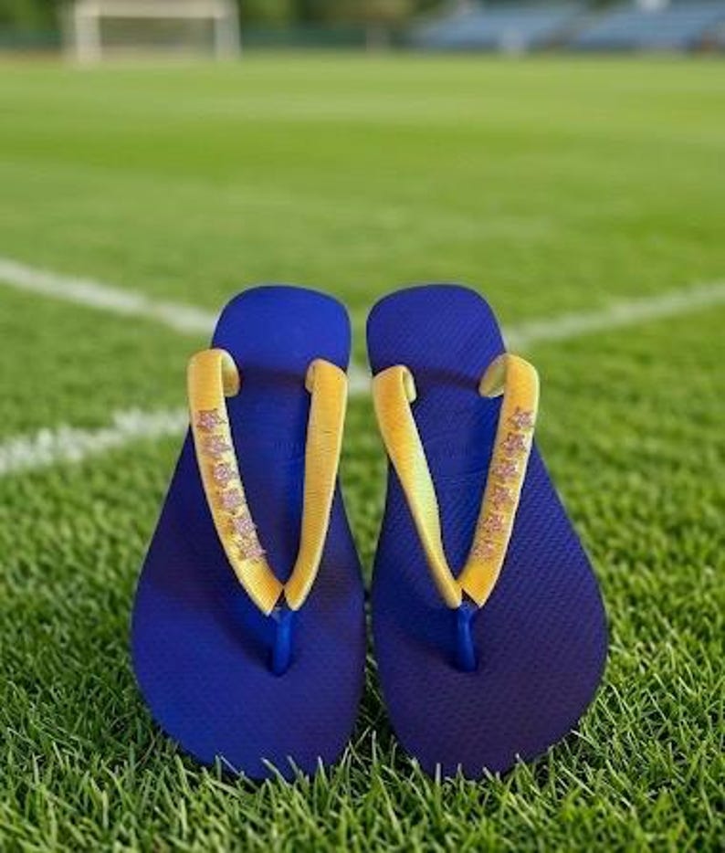 May include: A pair of blue flip-flops with yellow straps, resting on a green grassy field. The straps have a decorative pattern. The flip-flops are a casual footwear choice.