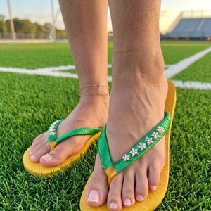 May include: Yellow flip-flops with green straps adorned with white flower-shaped embellishments. The flip-flops are worn on a green grassy surface.