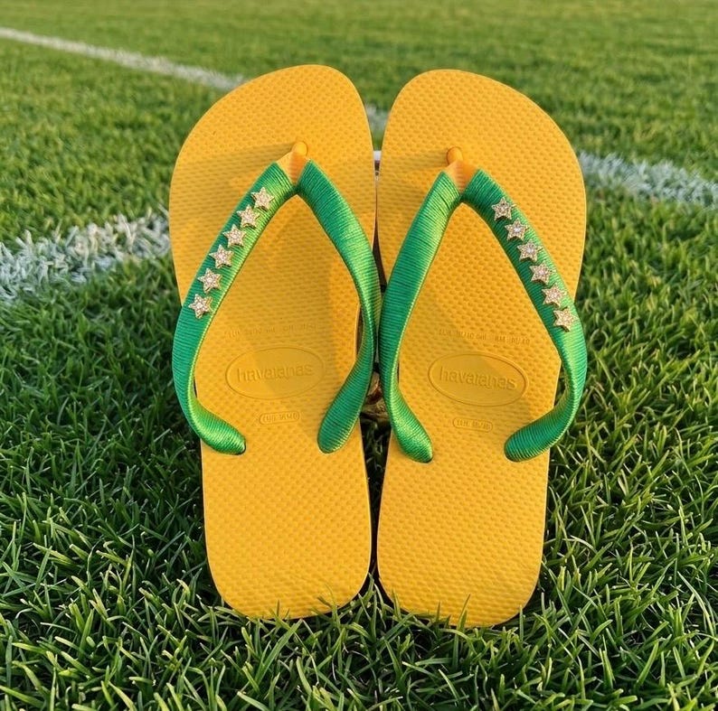 May include: A pair of yellow Havaianas flip-flops with green straps adorned with star-shaped embellishments. The flip-flops are resting on a green grassy surface, suggesting outdoor wear.