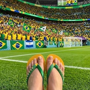 May include: Yellow and green flip-flops with star embellishments on the straps rest on a green field. In the background, a stadium is filled with fans waving Brazilian flags, creating a vibrant atmosphere.