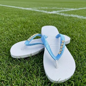 May include: White flip-flops with light blue straps adorned with small, gold-colored star-shaped embellishments. The flip-flops are resting on a bed of green grass, with a soccer field in the background.