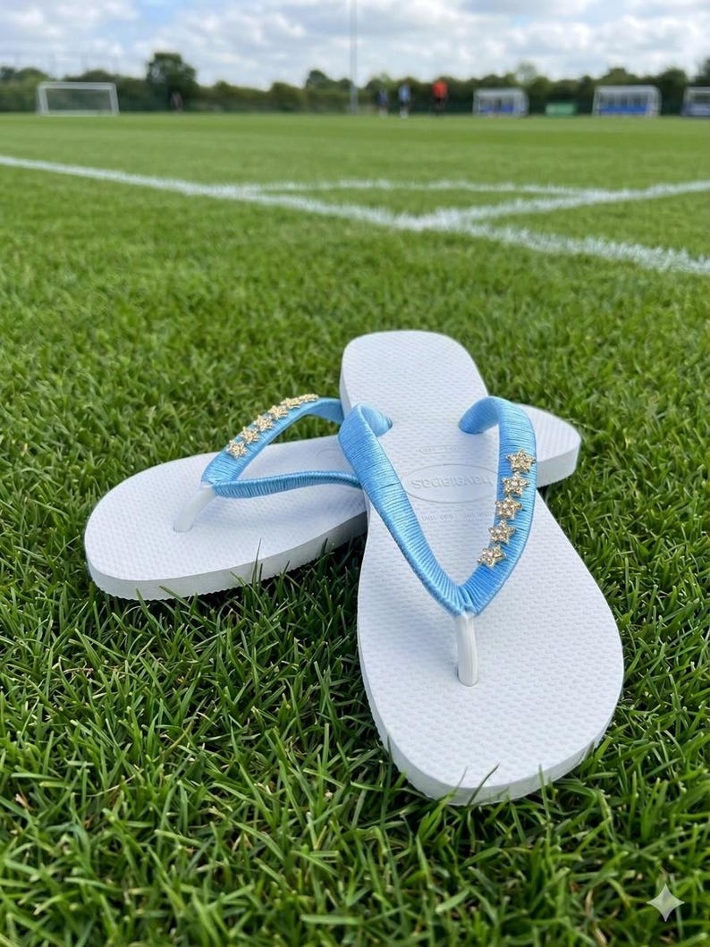 May include: White flip-flops with light blue straps adorned with small, gold-colored star-shaped embellishments. The flip-flops are resting on a bed of green grass, with a soccer field in the background.