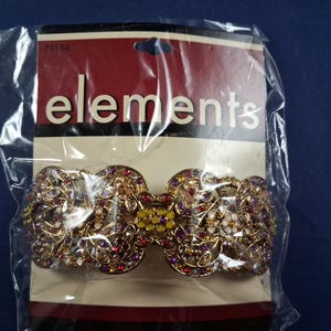 May include: A gold-toned hair clip with intricate floral designs, embellished with colorful rhinestones. The clip is packaged in a clear plastic bag with a cardboard backing that reads "elements".