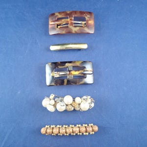 May include: Assortment of hair clips and barrettes on a blue background. Includes two tortoise shell rectangular clips, a gold-toned barrette, a pearl and rhinestone clip, a beaded clip, and a pink ribbed barrette.