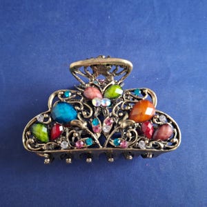 May include: Ornate, antique-style hair claw clip. The clip features a bronze-colored metal frame with intricate filigree designs. It is adorned with colorful, faceted gemstones in shades of blue, green, orange, and pink, adding a touch of sparkle.