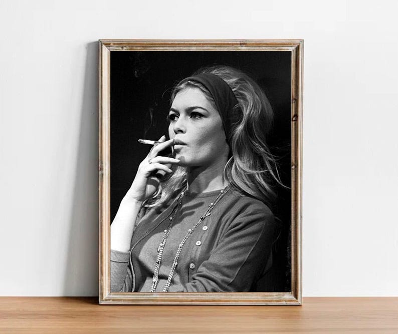 May include: Black and white framed portrait of a woman with long blonde hair, wearing a headband, cardigan, and necklace, smoking a cigarette. The image has a vintage aesthetic.