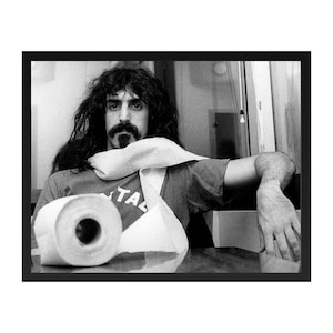 Puede incluir: Fotografía en blanco y negro enmarcada que presenta a un hombre con pelo largo y bigote, vistiendo una camiseta con la palabra "TAL". Un rollo de papel higiénico y un trozo de papel en primer plano.