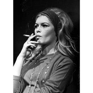 May include: Black and white print of a woman smoking a cigarette, wearing a headband and a long necklace. The print measures 24 x 36 inches. The woman is wearing a button-down cardigan. The print has a vintage aesthetic.