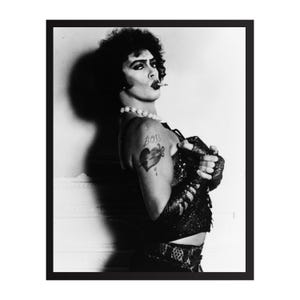 Peut inclure: Photographie en noir et blanc d'une personne en costume de scène, fumant une cigarette. La personne porte un collier de perles, un haut à paillettes et un tatouage avec l'inscription "Boss" et un motif de cœur et de poignard. L'image est encadrée d'un bord noir.