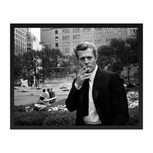 Puede incluir: Fotografía enmarcada en blanco y negro de un hombre con traje fumando un cigarrillo. El hombre está en un parque con una fuente y edificios al fondo. La imagen es un retrato.