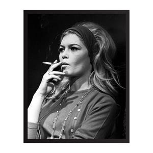 May include: Black and white portrait of a woman smoking a cigarette. She wears a dark headband, a button-down cardigan, and a long necklace. Her hair is styled in a bouffant, and she has hoop earrings. The image is framed in a black border.
