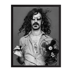 Puede incluir: Retrato en blanco y negro de un hombre con pelo largo y rizado, gafas de sol y bigote, sosteniendo un ventilador vintage y un cable de alimentación. El hombre lleva una camisa abotonada.