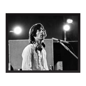 Puede incluir: Fotografía en blanco y negro de un hombre con pelo largo, camisa blanca y auriculares, cantando a un micrófono. La imagen es un plano medio, con luces de escenario en el fondo.