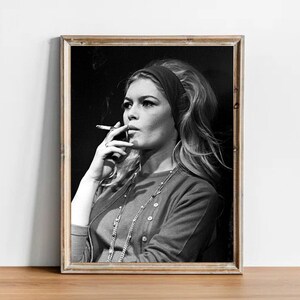 May include: Black and white framed portrait of a woman with long blonde hair, wearing a headband, cardigan, and necklace, smoking a cigarette. The image has a vintage aesthetic.