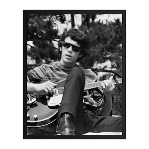 Puede incluir: Fotografía en blanco y negro de una persona con gafas de sol tocando una guitarra eléctrica. La persona lleva una camisa a rayas y pantalones negros. La guitarra es oscura con detalles plateados.