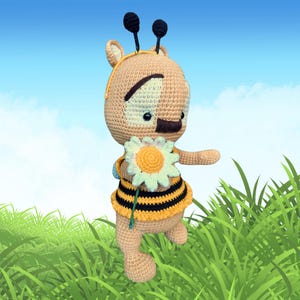 May include: A crocheted bee doll with a yellow and black striped outfit, holding a white and yellow daisy. The doll has a tan body, black antennae, and a sweet expression. The background features green grass and a blue sky.