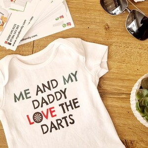 Op de afbeelding: Witte babybody met de tekst "ME AND MY DADDY LOVE THE DARTS" in zwart, groen en rood. De body ligt op een houten oppervlak met een zonnebril, een vetplant en visitekaartjes.