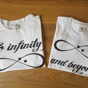 Könnte beinhalten: Zwei weiße T-Shirts mit schwarzem Text und Unendlichkeitszeichen-Designs. Ein Shirt sagt "To infinity" und das andere "and beyond". Jedes Shirt hat ein kleines Herzsymbol innerhalb des Unendlichkeitszeichens.