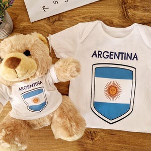 May include: A plush teddy bear wearing a white t-shirt with the word "ARGENTINA" and the Argentinian flag. A matching white baby onesie with the same design is also present. The items are on a wooden surface.