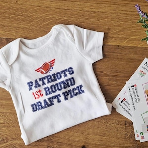 Op de afbeelding: Witte babybody met de tekst "PATRIOTS 1ST ROUND DRAFT PICK" in blauw en rood. De body heeft een football-afbeelding. Het kledingstuk is gemaakt van zachte stof en is ontworpen voor baby's.
