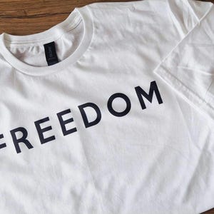 May include: White t-shirt with the word "FREEDOM" printed in bold black letters across the front. The shirt has a crew neck and short sleeves. The Gildan brand tag is visible at the neckline.