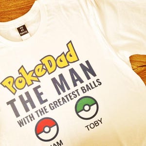 May include: White t-shirt with the text "PokeDad THE MAN WITH THE GREATEST BALLS". The design includes two Poke Ball graphics and the names "IAM" and "TOBY". The shirt is on a wooden surface.