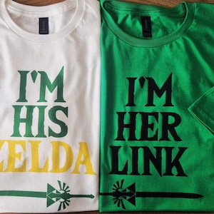 Può includere: Due magliette, una bianca e una verde, con testo e grafica abbinati. La maglietta bianca dice "I'M HIS ZELDA" in verde e giallo. La maglietta verde dice "I'M HER LINK" in nero. Entrambe hanno una grafica a spada.