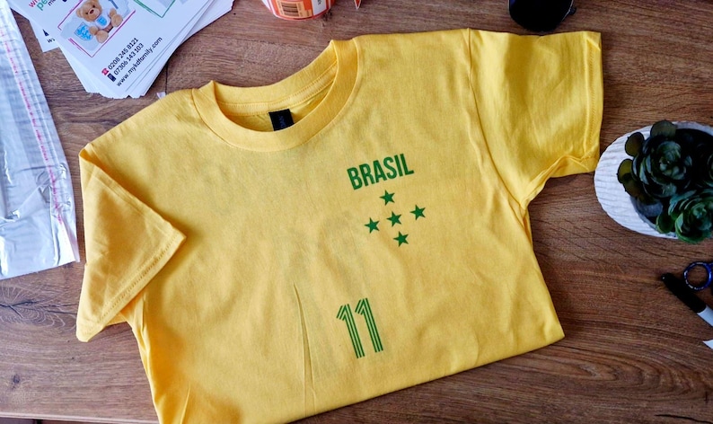 K&ouml;nnte beinhalten: Ein gelbes T-Shirt mit dem gr&uuml;nen Wort "BRASIL", f&uuml;nf gr&uuml;nen Sternen und der Nummer "11". Das Shirt besteht aus einem weichen Material und ist f&uuml;r den t&auml;glichen Gebrauch geeignet.