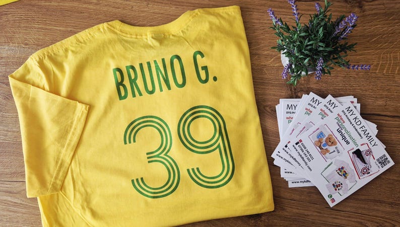 May include: Yellow t-shirt with the name "BRUNO G." and the number "39" printed in green. A small potted plant with purple flowers and business cards are also visible.