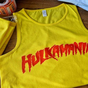 Puede incluir: Camiseta sin mangas amarilla con la palabra "HULKAMANIA" impresa en rojo. A la izquierda, una camiseta amarilla doblada con un gráfico rojo. Un rollo de etiquetas en el fondo.