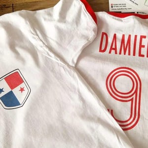 May include: Two white t-shirts with red trim. One shirt features a shield emblem with red, white, and blue sections and stars. The other shirt has the name "DAMIEN" and the number "9" in red.
