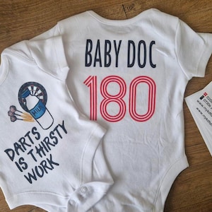 Op de afbeelding: Twee witte babyrompers. Eén heeft een afbeelding van een dartbord en een melkfles met de tekst "DARTS IS THIRSTY WORK". De andere zegt "BABY DOC 180" in rood en zwart. Een flyer ligt rechts.