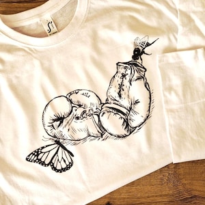 以下が含まれることがあります： ボクシンググローブ、蝶、蜂の白黒イラストが描かれた白いTシャツ。デザインは前面にプリントされています。Tシャツはクルーネックで半袖です。
