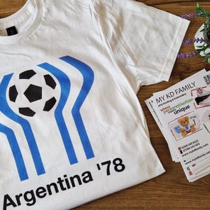 Puede incluir: Camiseta blanca con un diseño de balón de fútbol azul y negro y el texto "Argentina '78". La camiseta está sobre una superficie de madera con otros artículos, incluyendo tijeras y pegatinas.