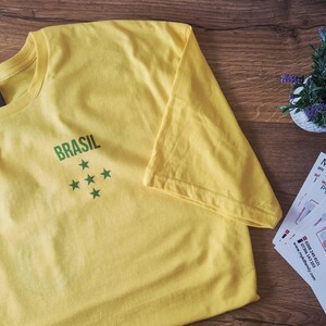 May include: A yellow t-shirt with the word "BRASIL" printed in green, and five green stars. The shirt is made of soft material and is folded on a wooden surface. A small plant and some cards are also visible.