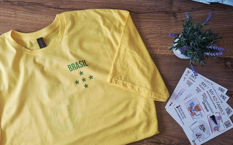 May include: A yellow t-shirt with the word "BRASIL" printed in green, and five green stars. The shirt is made of soft material and is folded on a wooden surface. A small plant and some cards are also visible.