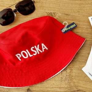Puede incluir: Un sombrero de pescador rojo con la palabra "POLSKA" en letras blancas. El sombrero tiene un ribete blanco y una etiqueta "reciclado". Gafas de sol están colocadas cerca. También se ven tarjetas de visita.