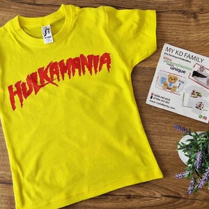 Puede incluir: Una camiseta amarilla de manga corta con el texto rojo "HULKAMANIA" en la parte delantera. Una pequeña etiqueta es visible en el cuello. Una tarjeta de visita y una pequeña planta con flores moradas también están en la imagen.