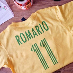 K&ouml;nnte beinhalten: Ein gelbes T-Shirt mit dem Namen "ROMARIO" und der Nummer "11" in Gr&uuml;n aufgedruckt. Das Shirt liegt flach auf einer Holzoberfl&auml;che, mit anderen Gegenst&auml;nden wie einer Klebebandrolle und bedruckten Papieren.