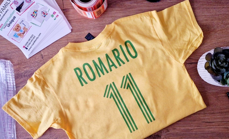 K&ouml;nnte beinhalten: Ein gelbes T-Shirt mit dem Namen "ROMARIO" und der Nummer "11" in Gr&uuml;n aufgedruckt. Das Shirt liegt flach auf einer Holzoberfl&auml;che, mit anderen Gegenst&auml;nden wie einer Klebebandrolle und bedruckten Papieren.