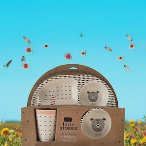 May include: A children's bamboo dinner set with a bear design, including a divided plate, bowl, small plate, cup, and spoons. The set is packaged in a brown box with the text "Little Stories". The background is a blue sky.