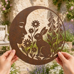 May include: A circular metal wall art piece with a rusted finish featuring a cutout design of a profile of a face, a rabbit, daisies, and foxglove flowers. The intricate silhouette is crafted from a single sheet of weathered brown metal.