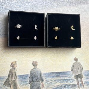 Op de afbeelding: Twee zwarte dozen met elk een set oorbellen met een hemels thema. Elke set bevat een planeet, een halve maan en een ster. De oorbellen zijn goudkleurig met sprankelende kristallen accenten. De achtergrond toont een potloodtekening van mensen op het strand.