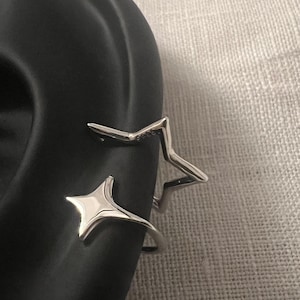 May include: Silver-coloured ear cuffs in a star shape. One is a solid star, the other an open design. The jewellery is displayed on a black surface. The cuffs are designed to be worn on the ear.