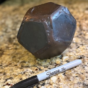 Puede incluir: Un objeto grande, de color marrón oscuro, multifacético, con forma geométrica. Un rotulador Sharpie negro y blanco de punta fina está al lado del objeto para comparar el tamaño. El objeto parece estar hecho de un material sólido.