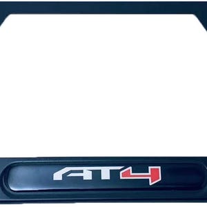 May include: Black license plate frame with the letters "AT4" in white and red. The frame has a rectangular shape with rounded corners and mounting holes. The "AT4" logo is centered on a black bar at the bottom of the frame.
