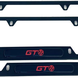 Könnte beinhalten: Zwei schwarze Kennzeichenrahmen mit rotem "GT"-Schriftzug und Logo. Die Rahmen sind rechteckig mit abgerundeten Ecken und Befestigungslöchern. Sie sind für Standard-Kennzeichen konzipiert.