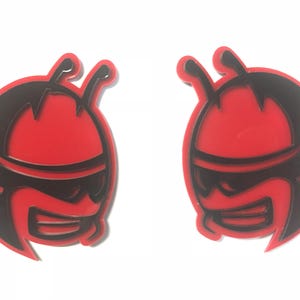 Puede incluir: Dos pegatinas rojas y negras con forma de insecto de dibujos animados. Cada pegatina presenta una forma ovalada roja con un contorno negro y una cara negra con gafas de sol. Las pegatinas tienen un acabado brillante y están sobre un fondo blanco.