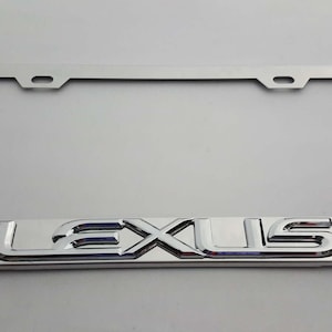 Op de afbeelding: Een chromen kentekenplaathouder met het woord "LEXUS" in reliëf in een vet, gestileerd lettertype. De houder is rechthoekig met afgeronde hoeken en montagegaten aan de bovenkant.